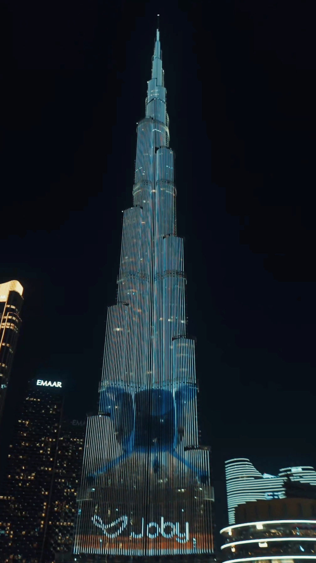 The Burj Khalifa illuminated at night with colorful projections, towering over nearby buildings.