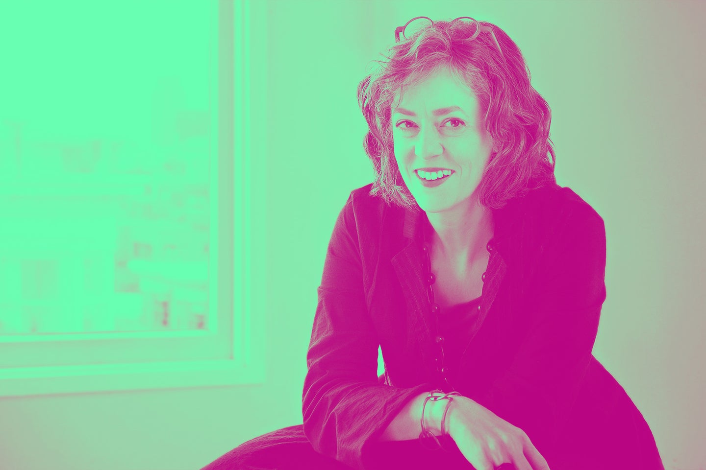A smiling woman with curly hair sitting near a window, wearing a black jacket and bracelets, in a vibrant, pink-tinted setting.