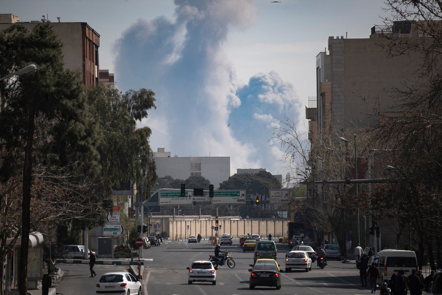 Smoke billows from buildings in the distance, viewed from a city street with vehicles and trees lining the road.