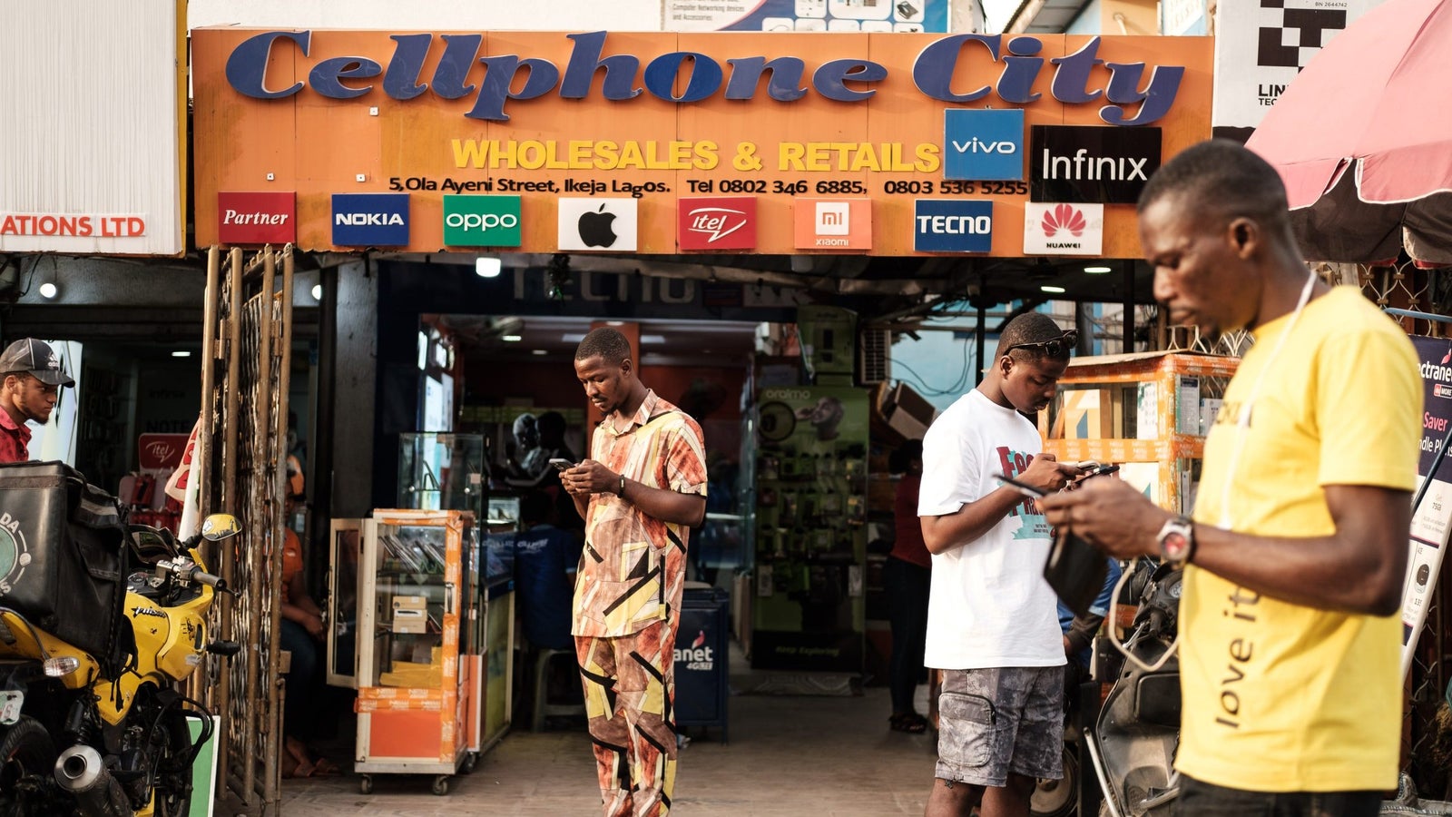 A storefront labeled "Cellphone City," with several men outside using their phones, surrounded by various mobile phone displays.