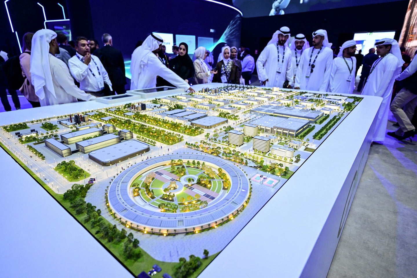 A detailed model of a futuristic city layout, with people observing and discussing around it at an exhibition.