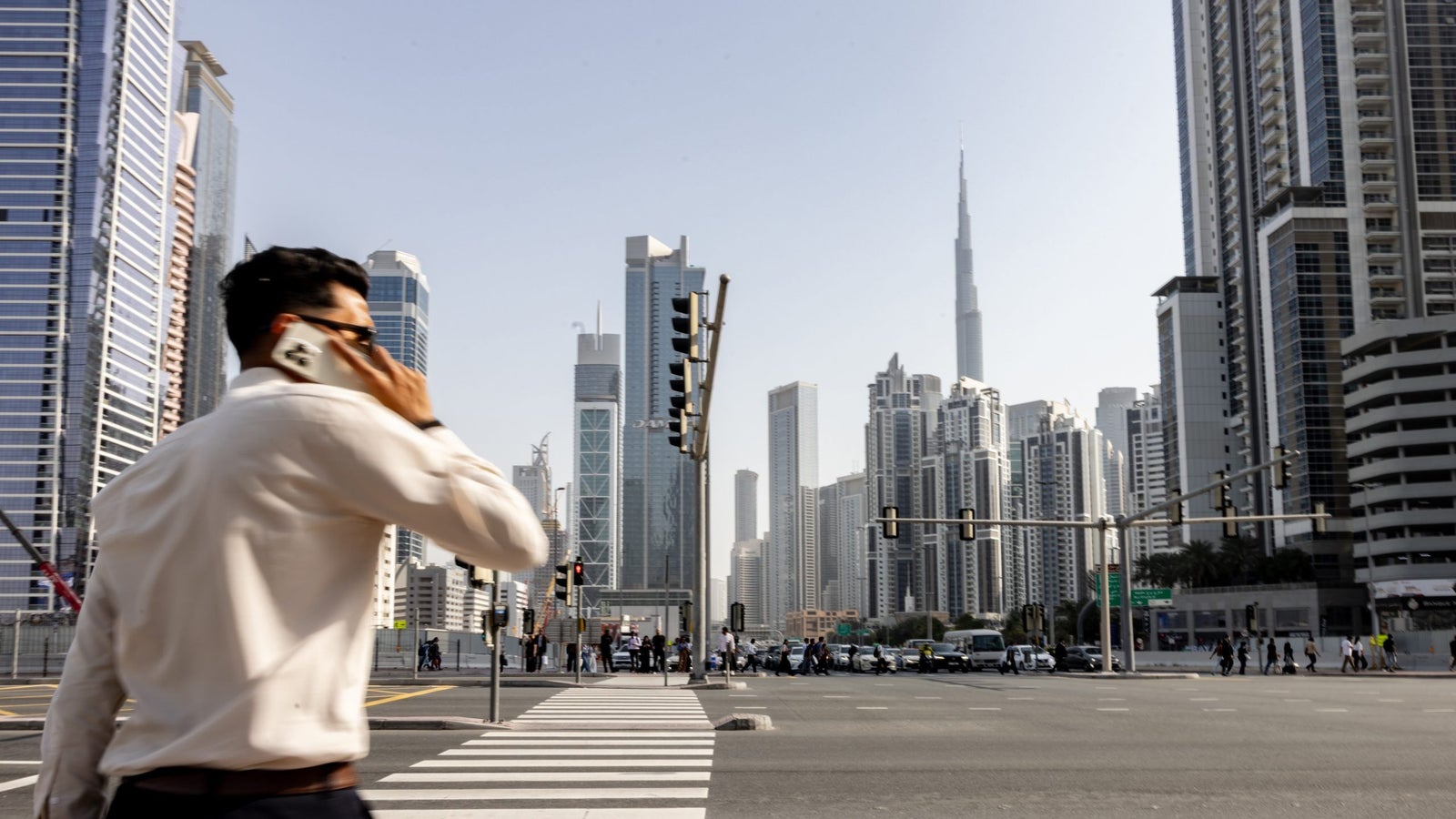 A businessman in a white shirt talks on a phone while crossing a busy street in Dubai, with tall skyscrapers in the background.