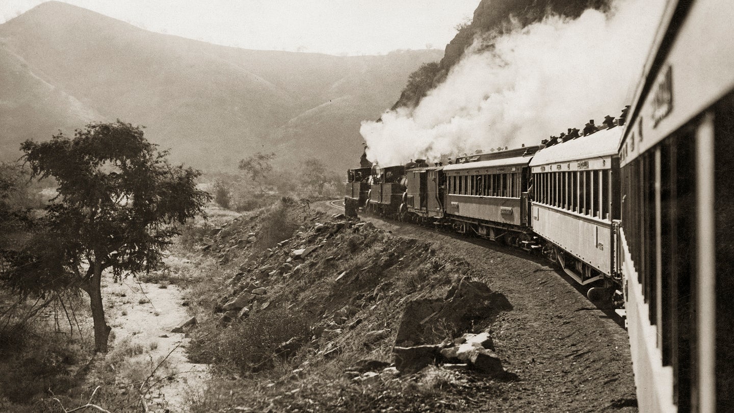 A vintage steam train travels along a winding track beside a rocky hillside, with smoke billowing from the engine.