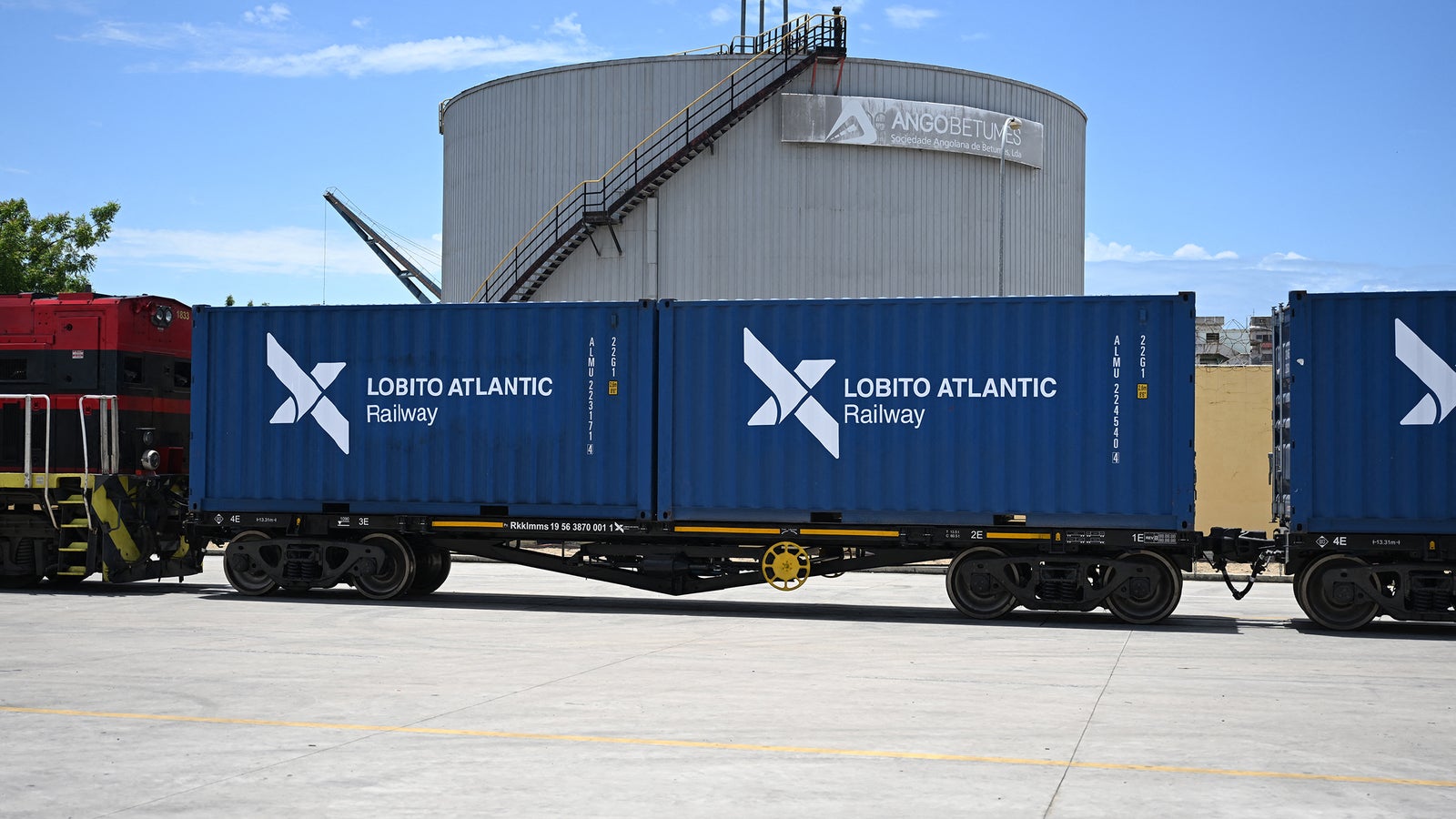 Blue shipping containers with "Lobito Atlantic Railway" label are parked next to a large storage tank under a clear sky.
