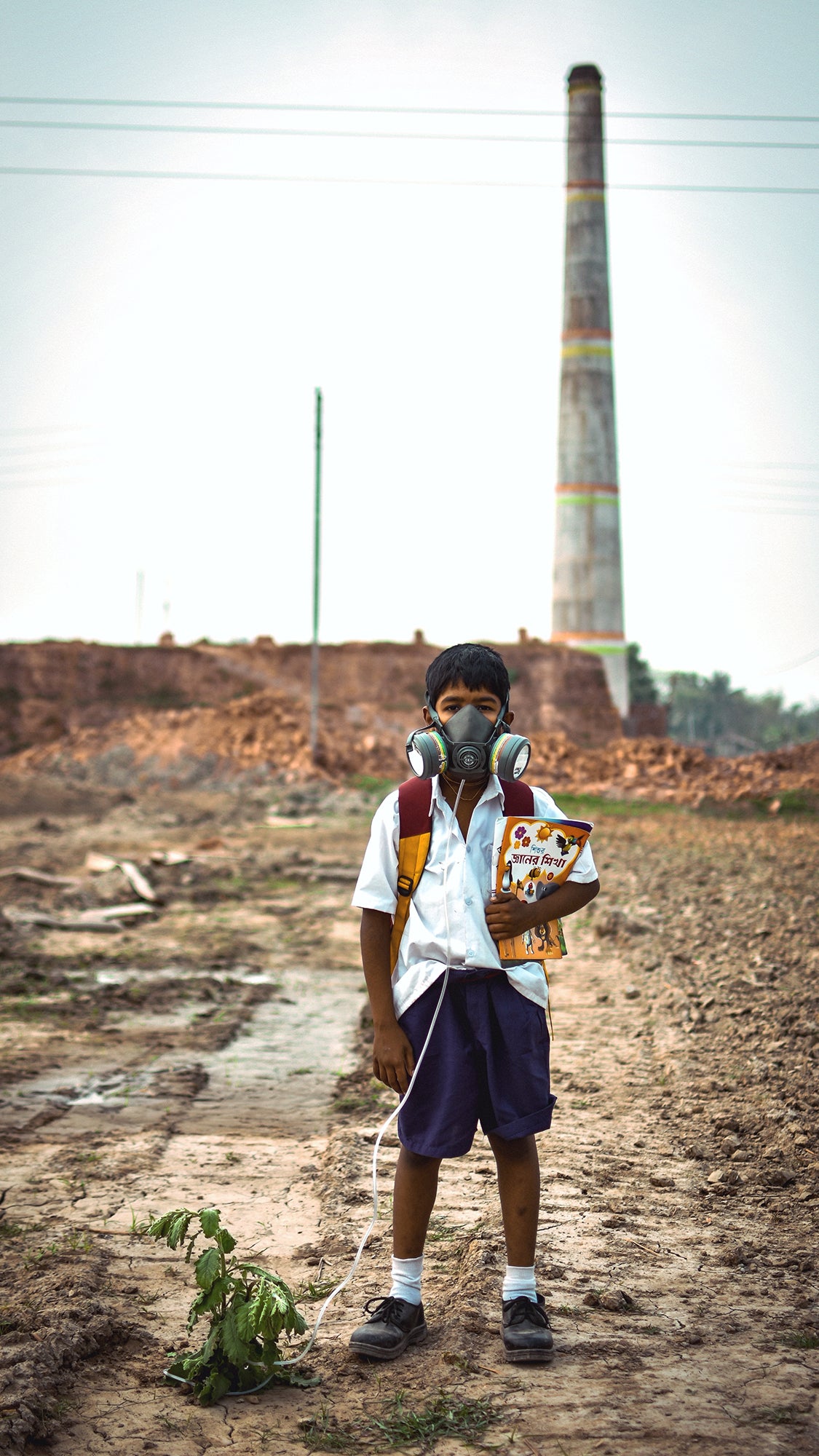 A schoolboy wearing a gas mask and headphones stands in a dusty, industrial area near a tall smokestack.