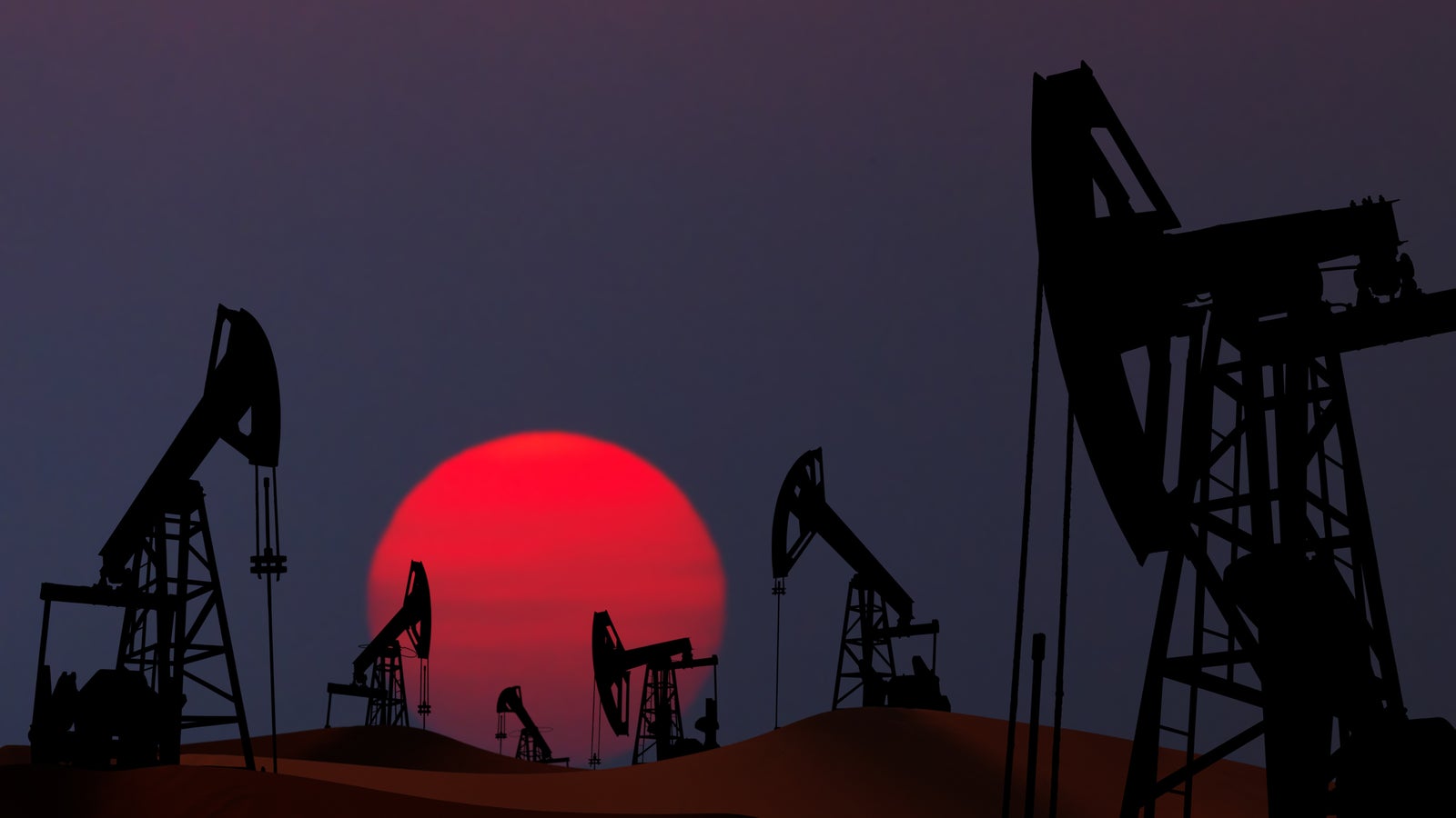 Silhouetted oil pumps against a large, vibrant red sunset, creating a stark contrast in the dusk sky.