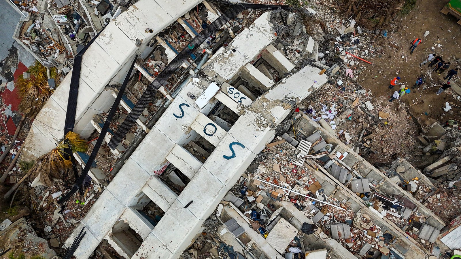 Aerial view of a collapsed building with the word "SOS" visible on a concrete slab, surrounded by debris and rescue workers