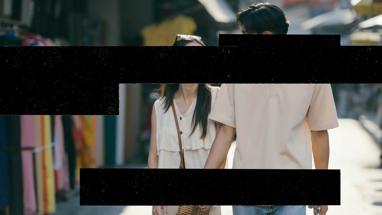 A couple, hand-in-hand, walks through a vibrant market street filled with colorful fabrics and soft sunlight