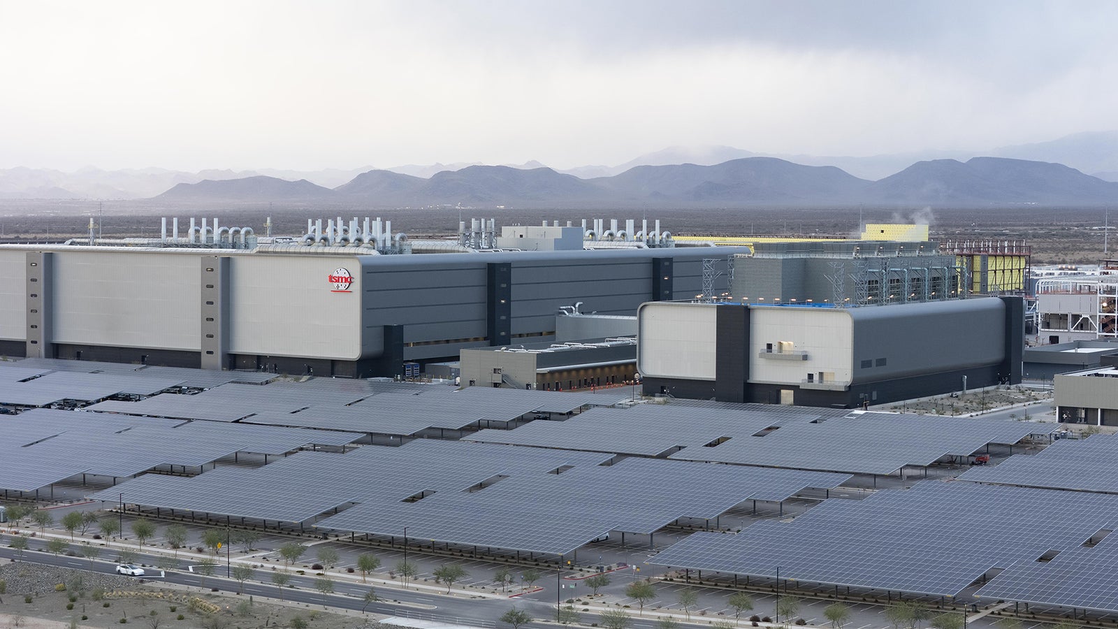 A large semiconductor manufacturing facility with solar panels in the foreground and mountains in the background under a cloudy sky