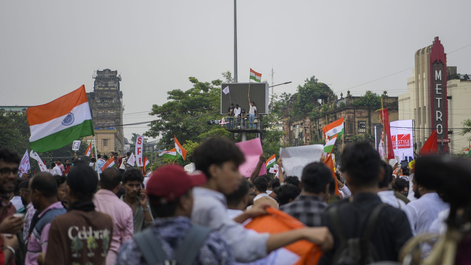 A large crowd gathers at a rally, waving flags, with a group on a platform and buildings in the background