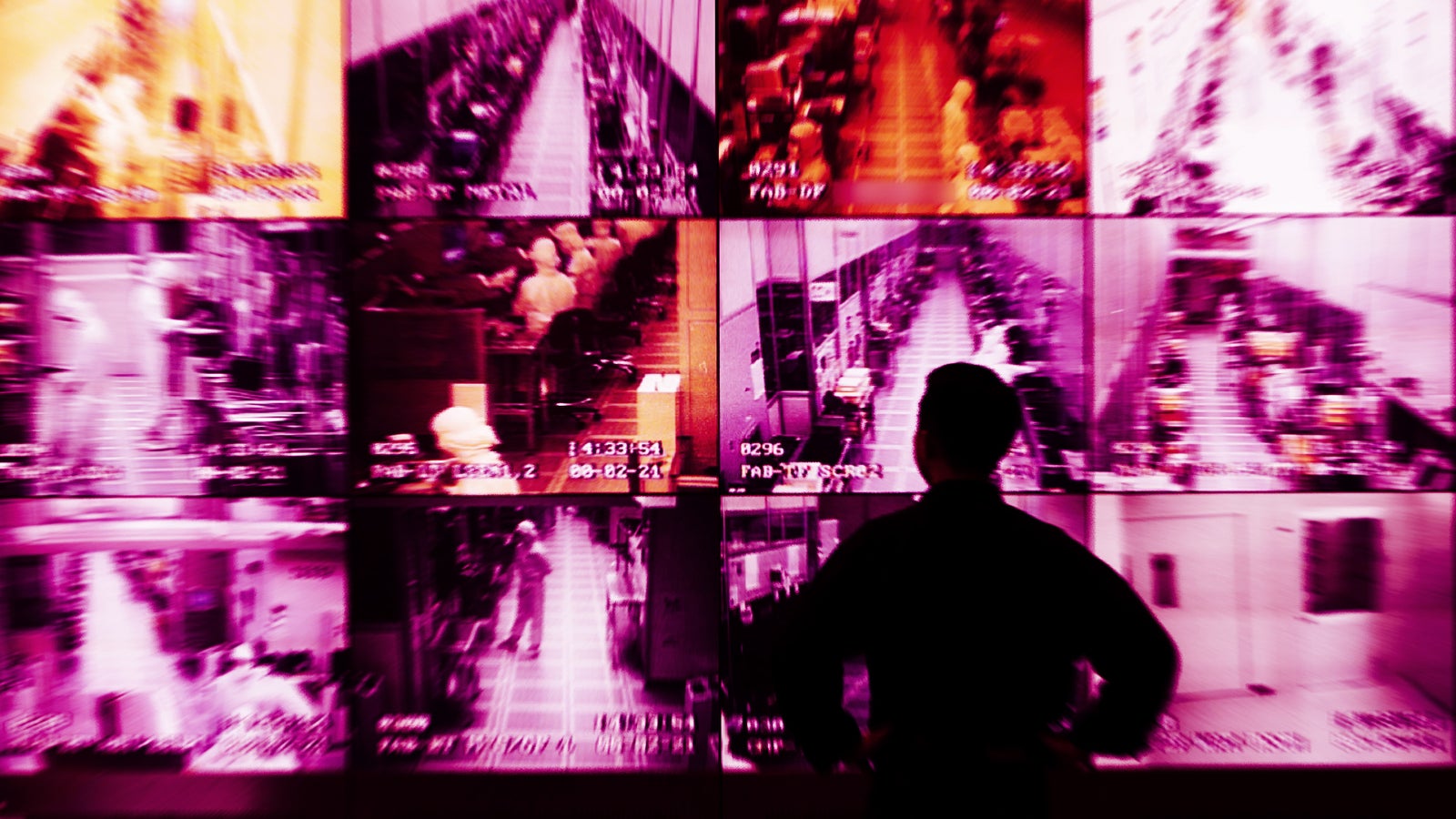 A person stands facing multiple surveillance monitors displaying footage in shades of pink and purple