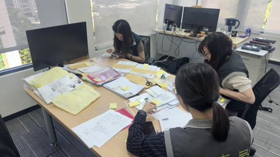 Three women work at a table covered with documents and stationery in a bright office space, focused on their tasks