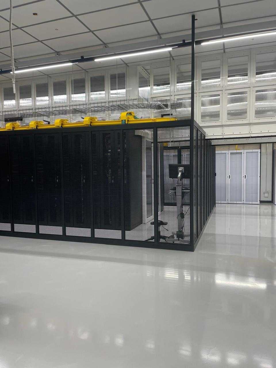 A spacious server room featuring rows of black mesh server racks, bright lighting, and a clean white floor