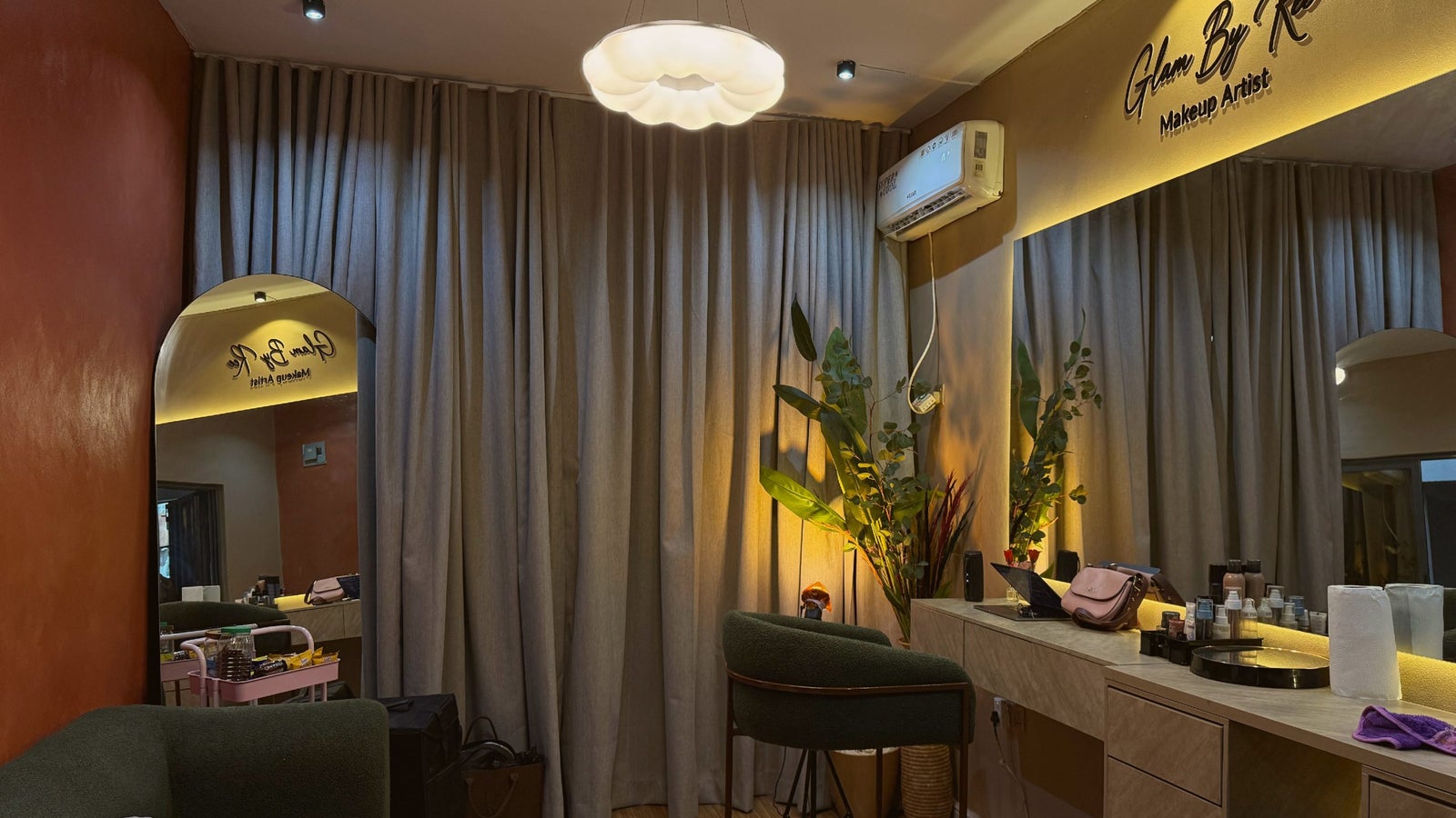 A makeup artist studio featuring a green chair, a vanity with beauty products, and a large mirror reflecting curtains
